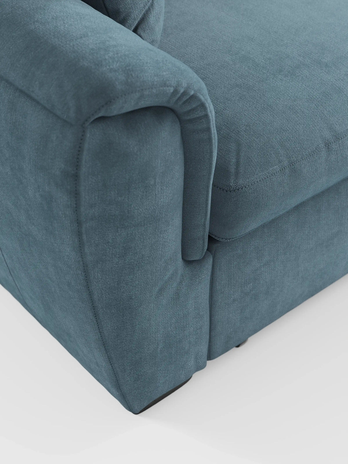 Primo piano dell'angolo e del bracciolo di un divano letto blu imbottito Titano con penisola reversibile, contenitore, poggiatesta reclinabili ed elemento estraibile in tessuto smacchiabile, firmato divitt.sbs. Il bracciolo presenta sottili dettagli di cucitura e curve morbide, che rispecchiano il design italiano contemporaneo. I morbidi cuscini di seduta esaltano il comfort, rendendo questo divano letto con chaise longue un elemento centrale e accogliente per qualsiasi soggiorno. Lo sfondo è costituito