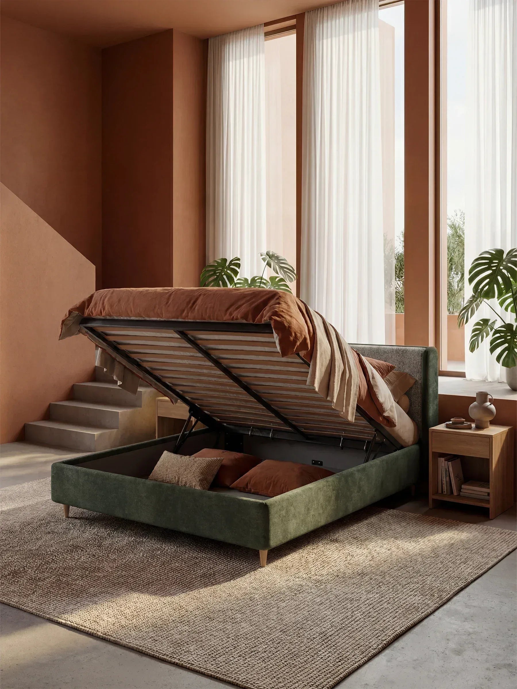 Una camera da letto moderna con il letto imbottito bicolore VALKIRIA con contenitore di Letti.store, biancheria da letto color ruggine, finestre a tutta altezza con tende trasparenti, piante rigogliose, un comodino e un tappeto beige su pavimenti in cemento inondati da una calda luce naturale.