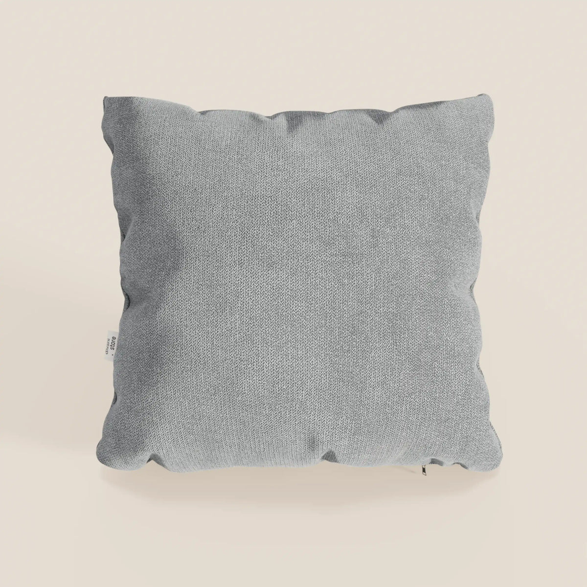 Il cuscino Destiny di divitt.sbs, 45x45 cm in morbido tessuto impermeabile grigio chiaro T02, ha una forma quadrata con angoli leggermente arrotondati e una piccola etichetta bianca sulla cucitura sinistra, su sfondo bianco sporco.