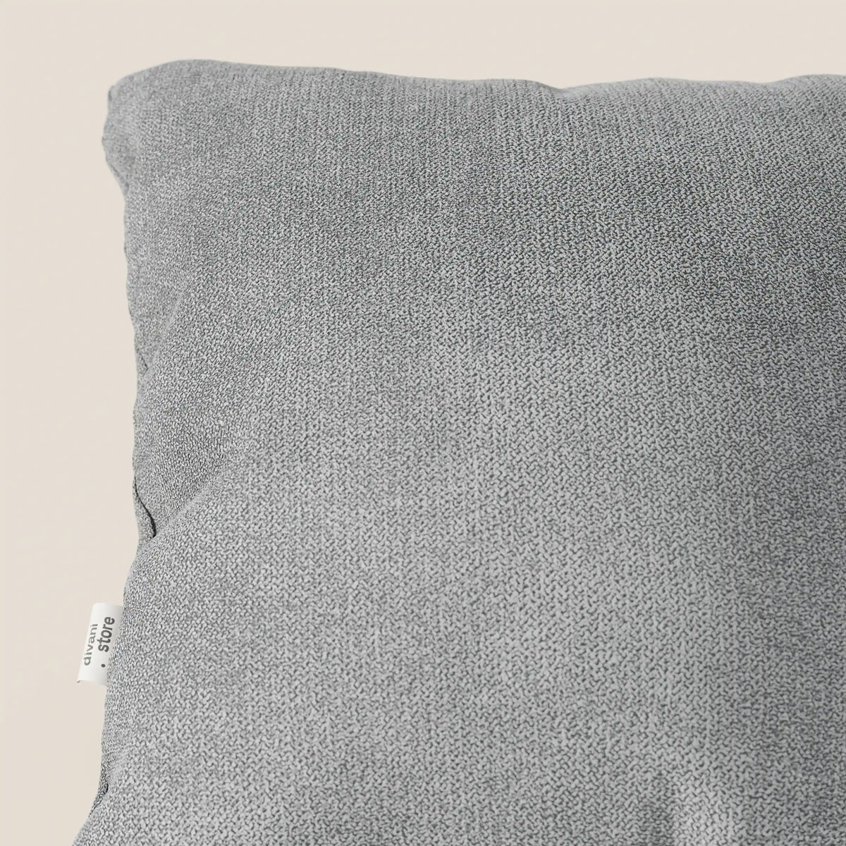Un primo piano del cuscino lavabile Destiny di divitt.sbs in tessuto morbido impermeabile T02 grigio chiaro, 45x45 cm, mostra la sua sottile trama a maglia e l'etichetta in tessuto bianco a sinistra, su uno sfondo beige chiaro.