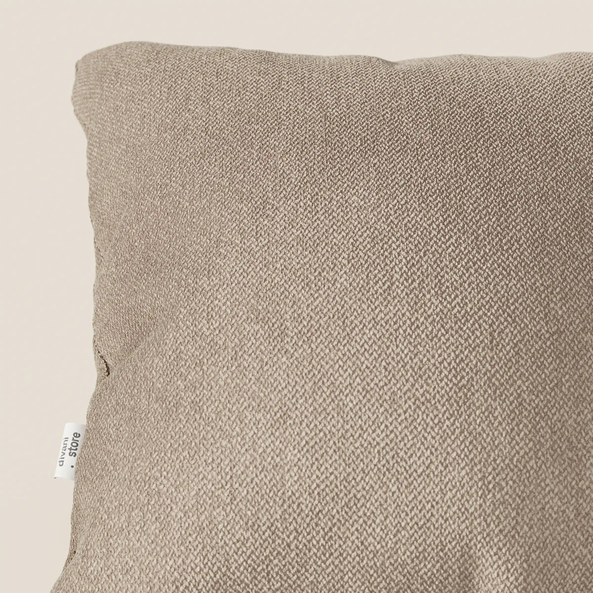 Primo piano dell'angolo superiore di un cuscino d'arredo beige Sogno di divitt.sbs in morbido tessuto smacchiabile T05, che mostra la trama a spina di pesce e un'etichetta bianca cucita. Lo sfondo è semplice e di colore chiaro.