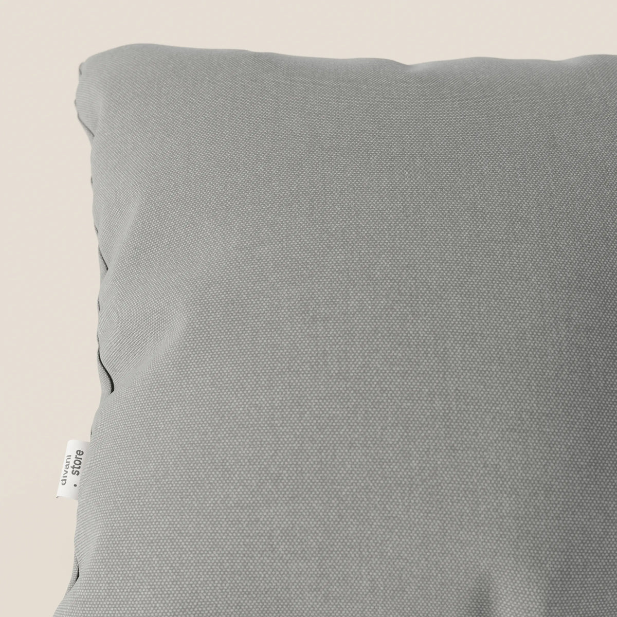 Primo piano del cuscino Honey 45x45 cm di divitt.sbs in T13 grigio tessuto simil cotone impermeabile, mostrato in diagonale con l'angolo in alto a sinistra, il lato e il cartellino in tessuto bianco su uno sfondo beige tenue.