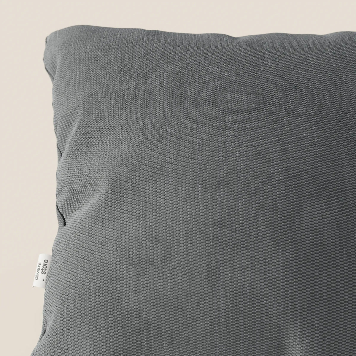 Primo piano del cuscino Miami di divitt.sbs in morbido tessuto impermeabile T14 grigio, che mostra l'angolo superiore sinistro con una piccola etichetta bianca sulla cucitura. Lo sfondo è un semplice beige chiaro.