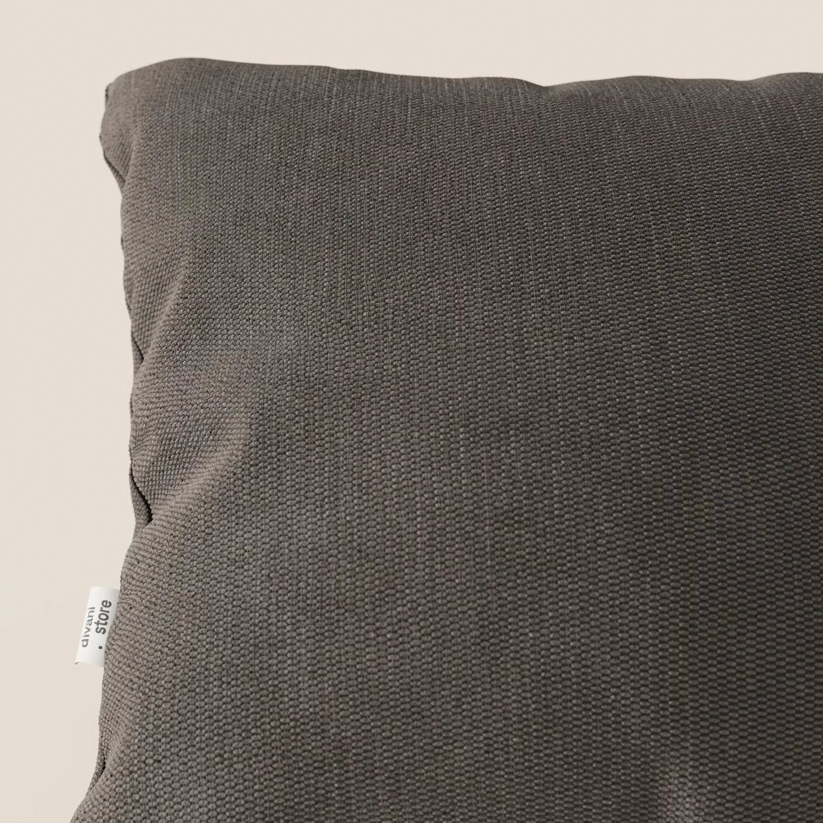 Un primo piano del cuscino Miami 45x45 cm di divitt.sbs in T14 marrone mostra il suo tessuto morbido e resistente all'acqua e la sua sottile trama. Una piccola etichetta bianca con testo nero è visibile sulla cucitura sinistra su uno sfondo beige.