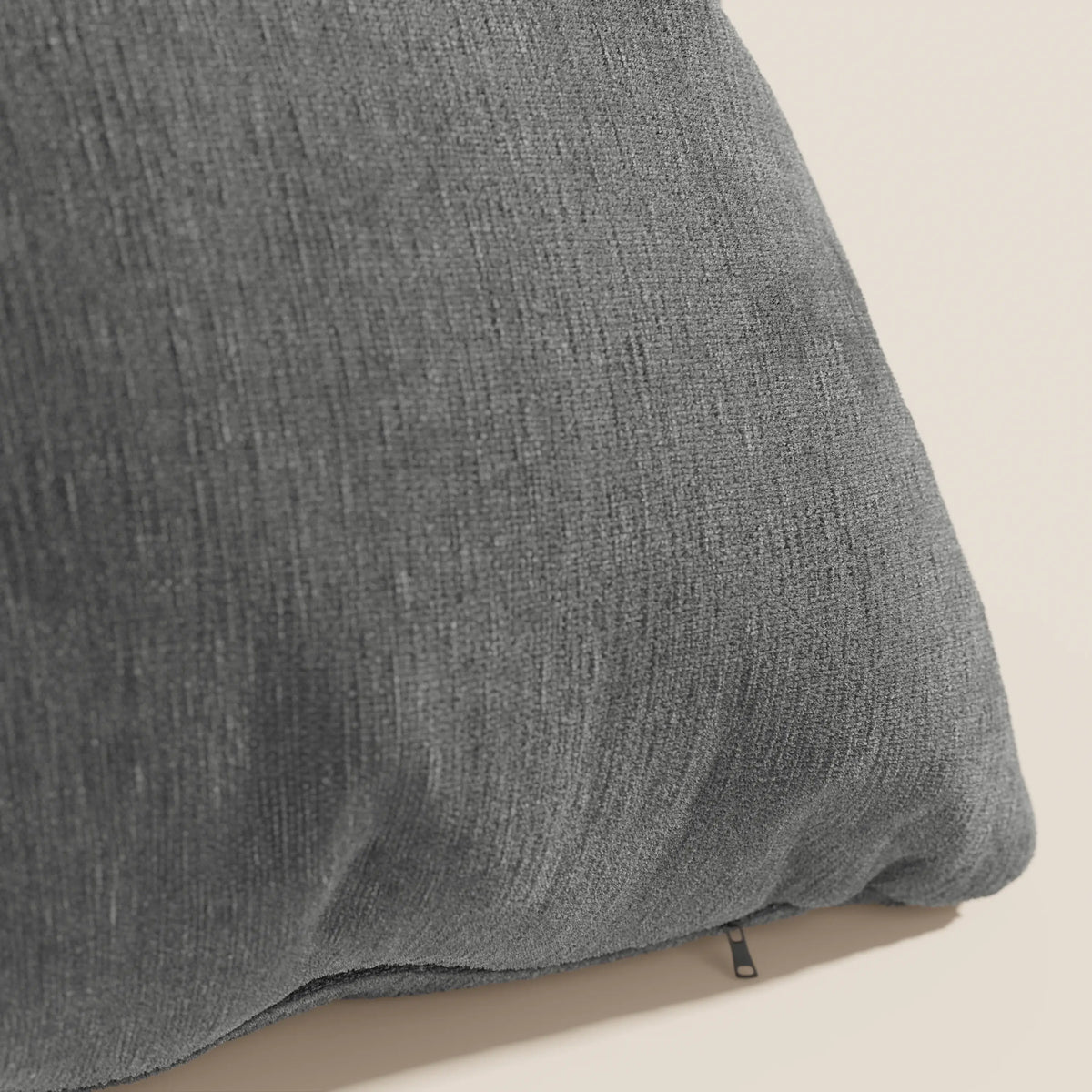 Un primo piano del cuscino Bolero di divitt.sbs in morbido tessuto grigio scuro T31 antimacchia, impermeabile e non restringente con cerniera inferiore a vista, esposto su uno sfondo beige chiaro per un look accogliente e contemporaneo.