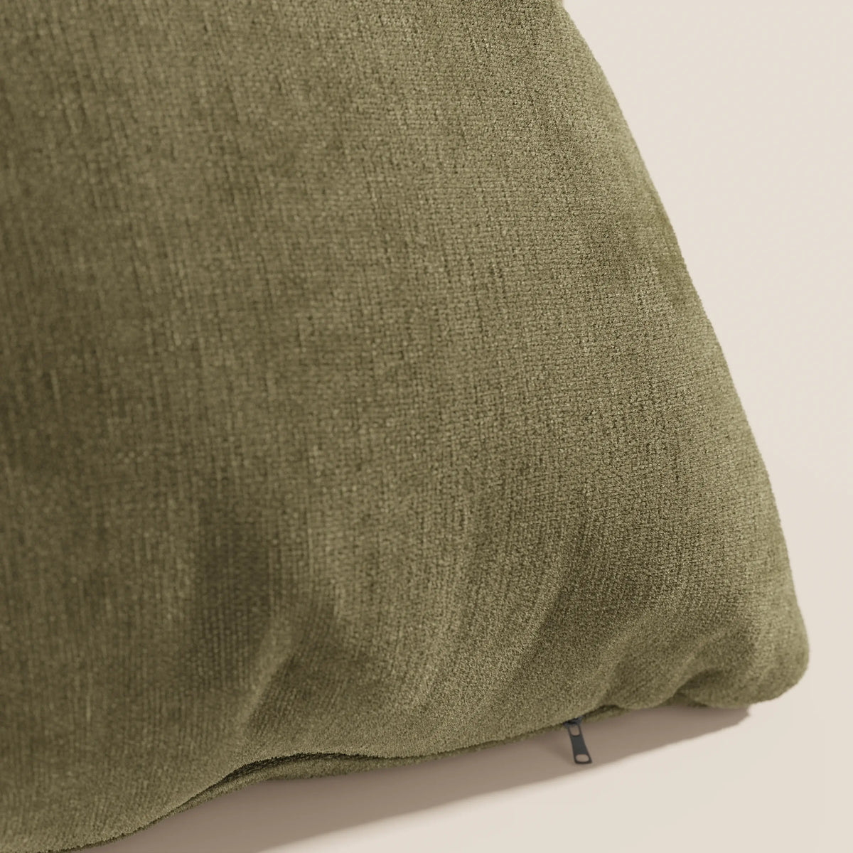Primo piano del cuscino Bolero di divitt.sbs in verde oliva, caratterizzato da un morbido tessuto T31 antimacchia, impermeabile e non restringente. In basso è visibile un piccolo tiretto nero per la chiusura lampo. Lo sfondo è beige chiaro.