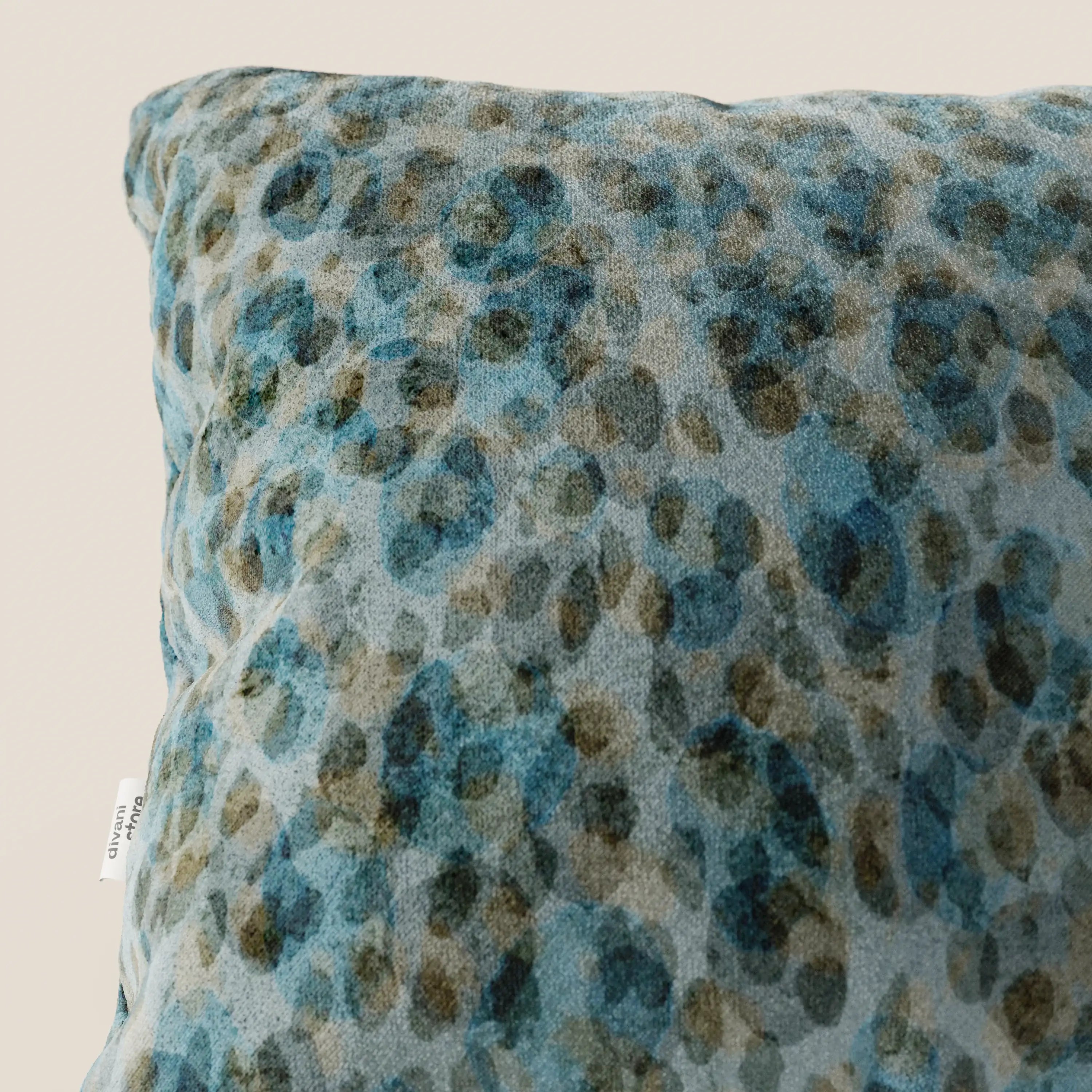 Primo piano del cuscino Brush 45x45 cm di divitt.sbs in morbido velluto blu impermeabile, caratterizzato da un motivo maculato acquerellato e da una piccola etichetta bianca con testo nero parzialmente visibile sul bordo.