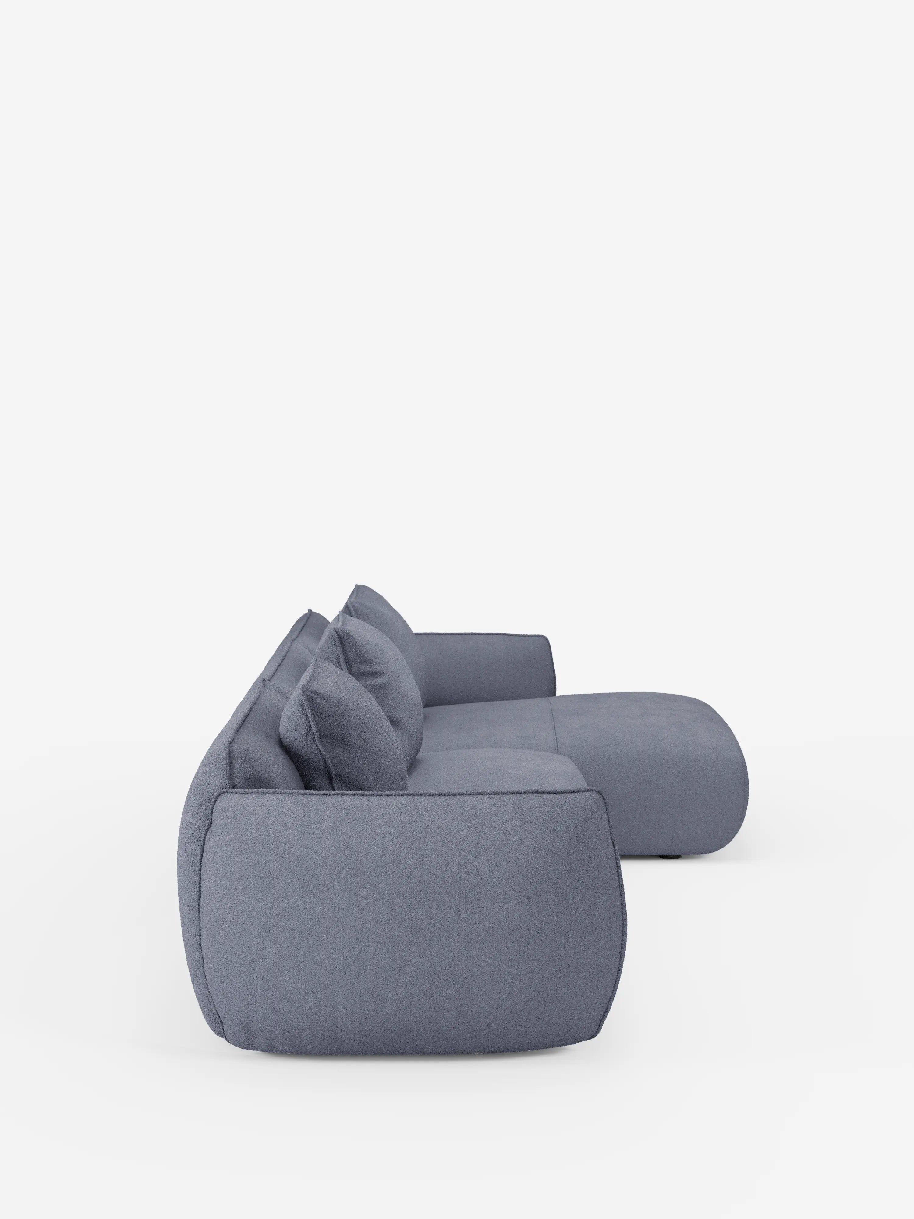 Vista laterale del divano angolare con penisola componibile Nimbus di divitt.sbs, rivestito in morbido tessuto T33 effetto cotone, resistente alle macchie, in grigio chiaro. Questo moderno divano componibile presenta cuscini soffici, bordi delicatamente arrotondati e due cuscini dello schienale abbinati, il tutto su uno sfondo bianco a tinta unita. Il design contemporaneo è sobrio e minimalista, e incarna al contempo comfort e stile.