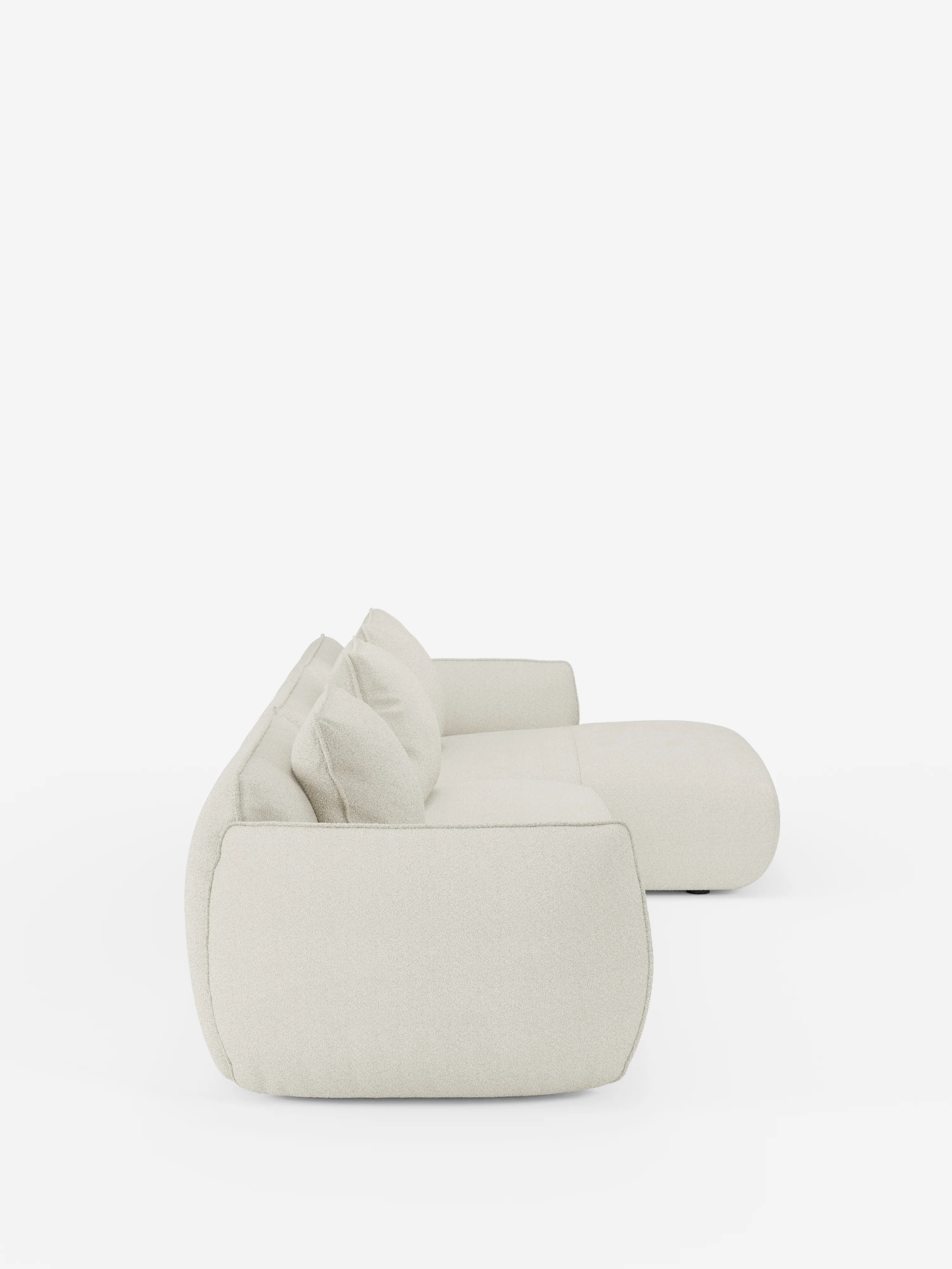 Vista laterale del divano angolare con penisola componibile Nimbus in morbido tessuto effetto cotone smacchiabile T33 di divitt.sbs, un divano moderno e minimalista color bianco sporco con bordi arrotondati e morbidi cuscini che risaltano su uno sfondo bianco a tinta unita. Questo modello contemporaneo presenta un profilo basso, due cuscini dello schienale e un ampio bracciolo per un maggiore comfort.