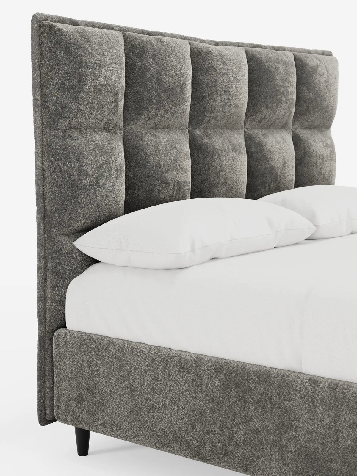 Un primo piano di un letto imbottito AURO con contenitore e testiera alta capitonné in ciniglia Oeko-Tex®, caratterizzato da una testiera alta in velluto grigio a motivi quadrati, lenzuola bianche e due morbidi cuscini su uno sfondo chiaro.