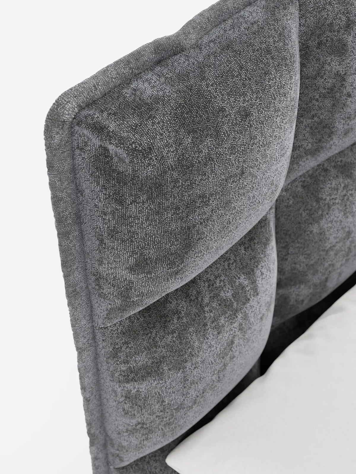 Primo piano della testiera capitonné grigio antracite di un letto imbottito AURO con contenitore e testiera alta in ciniglia Oeko-Tex®, che ne mette in risalto la morbida trama del tessuto sopra un angolo di un lenzuolo bianco su uno sfondo bianco.