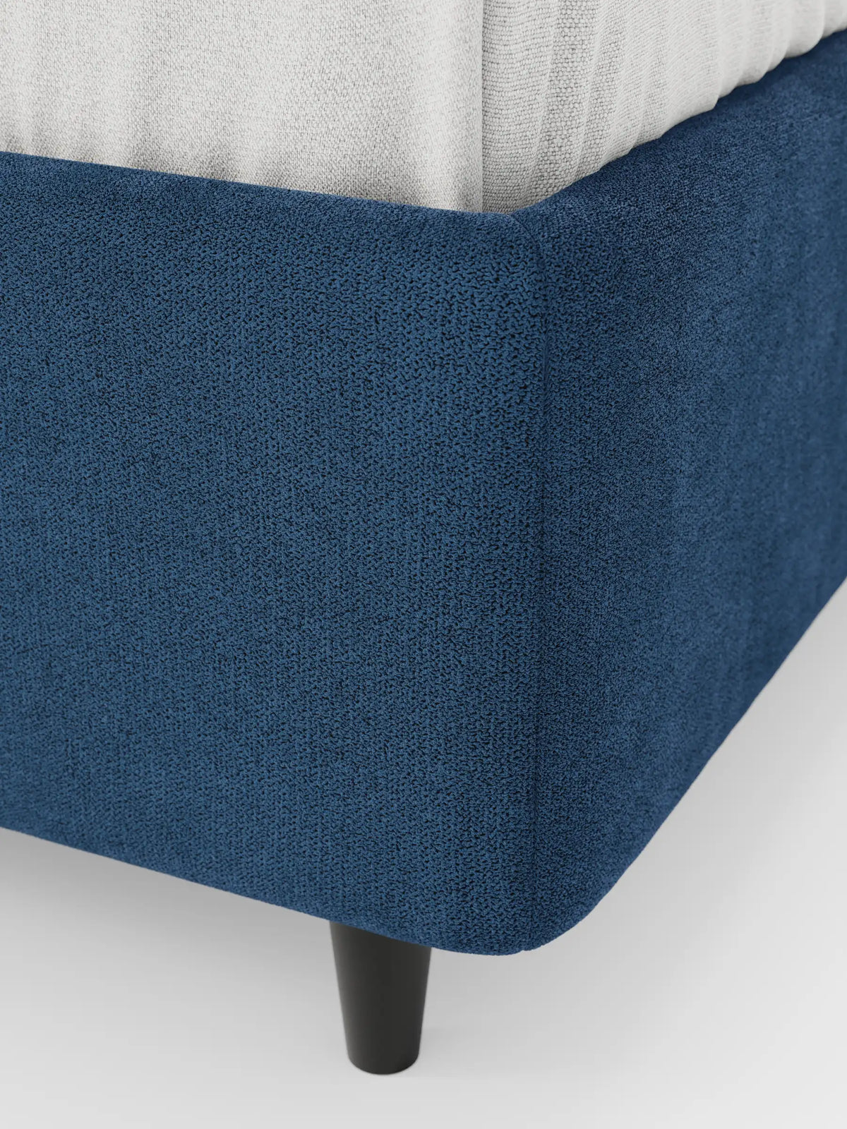 Un primo piano del letto sommier imbottito ERON con contenitore, realizzato in tessuto impermeabile certificato Oeko-Tex, caratterizzato da un rivestimento blu scuro, un angolo arrotondato su una gamba affusolata scura e una porzione di materasso bianco su uno sfondo a tinta unita.