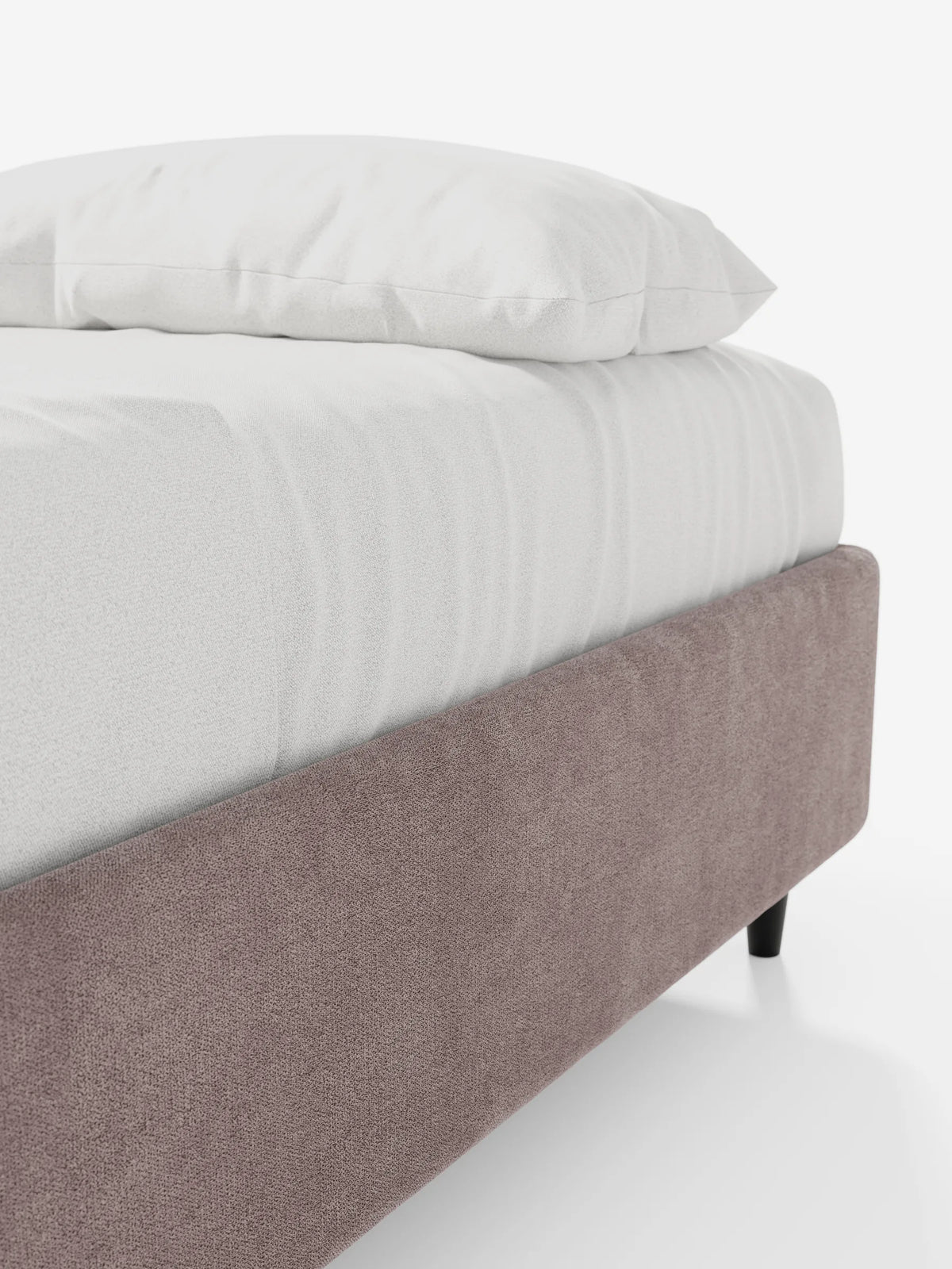 Vista angolare di un letto sommier imbottito ERON con vano contenitore in tessuto impermeabile certificato Oeko-Tex, caratterizzato da una base imbottita con finitura testurizzata color malva e da gambe corte scure, con biancheria da letto bianca su uno sfondo chiaro.