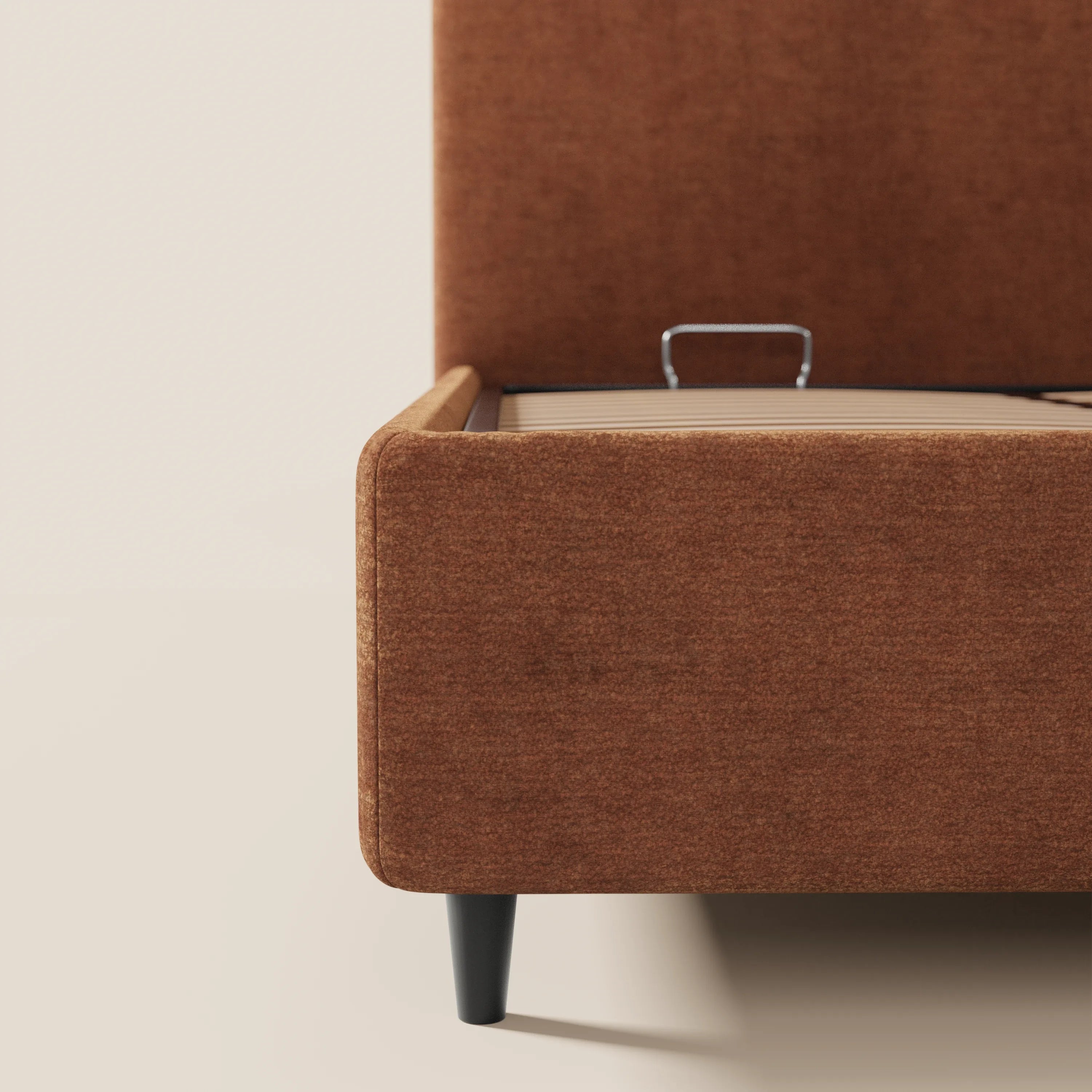 Un primo piano del letto KAELOS minimal imbottito di Letti.store ne mette in risalto il tessuto in ciniglia, le alte gambe nere, il sistema di fissaggio del materasso in metallo e la base in legno con vano contenitore e testiera arrotondata, il tutto su uno sfondo neutro.