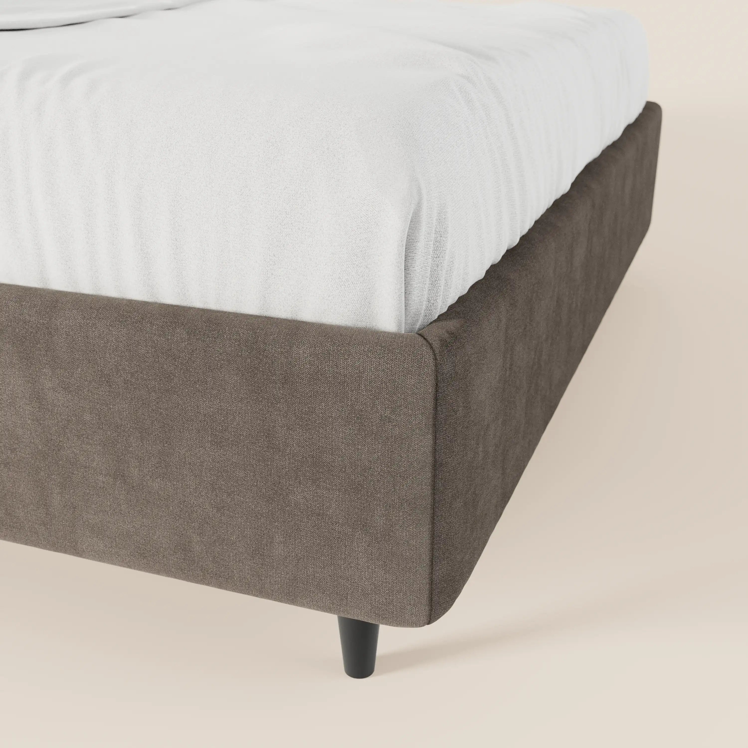 Un primo piano del letto sommier imbottito ERON di Letti.store ne mette in risalto la morbida struttura in tessuto impermeabile marrone, certificato Oeko-Tex, con gambe corte nere e un lenzuolo con angoli bianco, il tutto su uno sfondo beige chiaro a tinta unita.