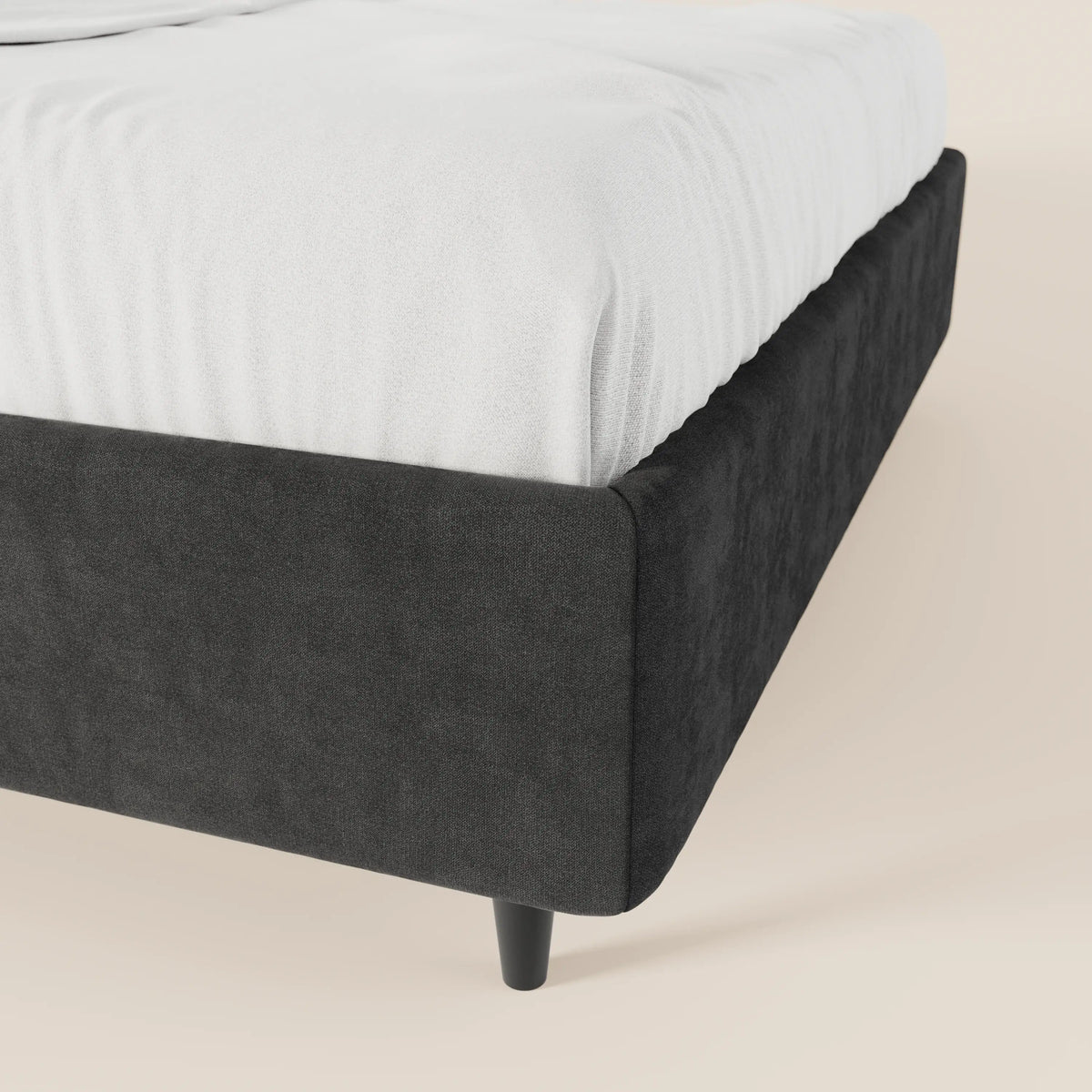 Un primo piano del letto sommier imbottito ERON di Letti.store, caratterizzato da un tessuto impermeabile nero certificato Oeko-Tex, gambe nere affusolate, senza testiera, un lenzuolo con angoli bianco e un pavimento beige chiaro, per uno stile pulito e minimalista.