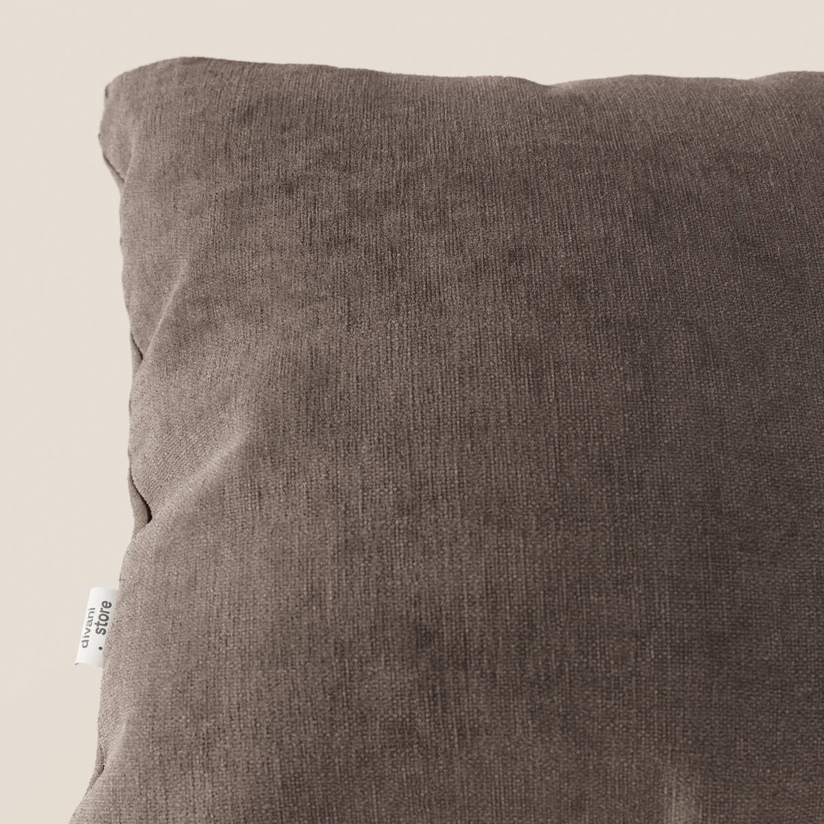 Primo piano di Bolero cuscino di divitt.sbs, in tessuto T31 marrone scuro, morbido, antimacchia, impermeabile e non restringente. L'immagine mostra l'angolo superiore sinistro con l'etichetta bianca su uno sfondo beige chiaro.