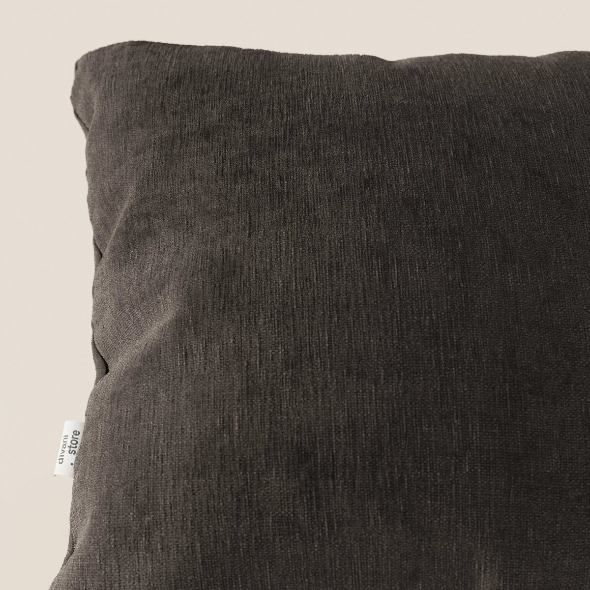 Primo piano del cuscino Bolero di divitt.sbs in tessuto T31 grigio scuro, smacchiabile, impermeabile e irrestringibile. Il cuscino, dalla consistenza morbida, è mostrato in posizione obliqua su uno sfondo beige chiaro, con un'etichetta bianca visibile sul bordo sinistro.