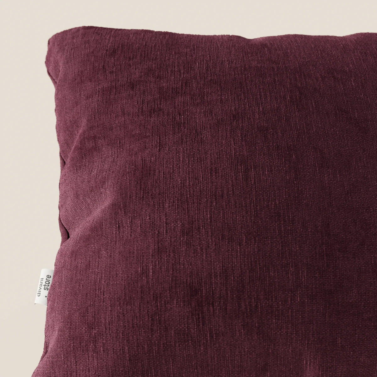 Primo piano del cuscino Bolero di divitt.sbs, un cuscino quadrato in morbido tessuto T31 antimacchia, impermeabile e antirestringimento. Il cuscino, di colore bordeaux scuro, presenta una piccola etichetta bianca sulla sinistra ed è inserito in uno sfondo beige semplice.