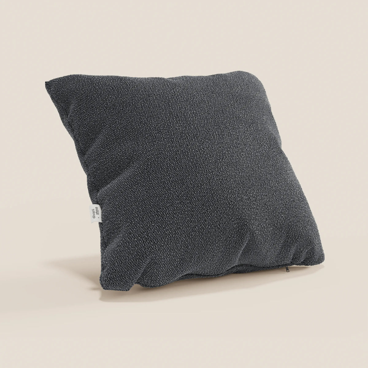Un cuscino Lambada grigio scuro di divitt.sbs, realizzato in tessuto fresco, antimacchia e impermeabile tipo cotone T33, con una piccola etichetta bianca su un lato, poggia morbidamente su una superficie beige chiaro su uno sfondo neutro.
