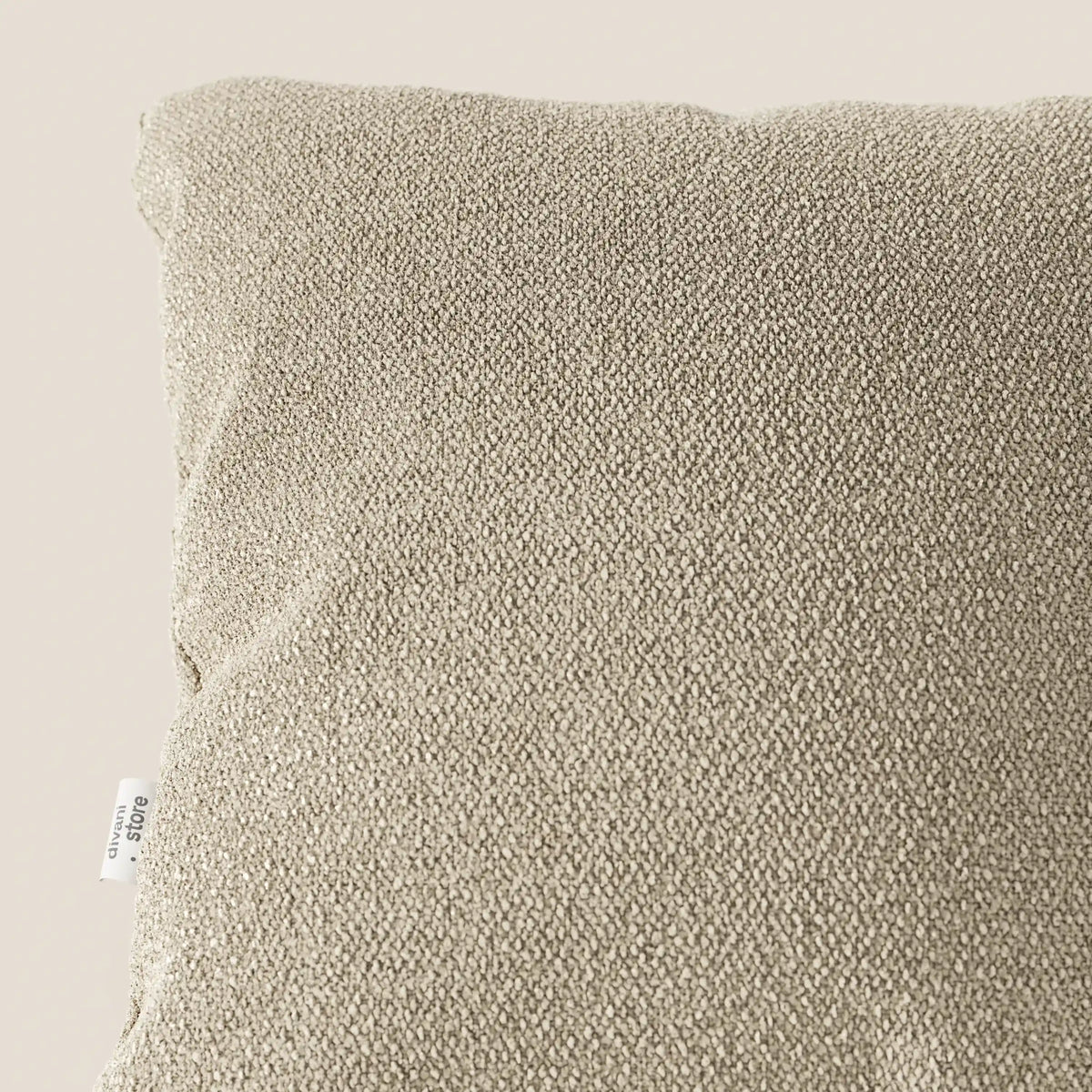 Primo piano del cuscino Lambada di divitt.sbs in beige, realizzato in tessuto T33 fresco, antimacchia e impermeabile, simile al cotone. La consistenza morbida e la piccola etichetta bianca sul bordo sinistro sono visibili su uno sfondo chiaro.