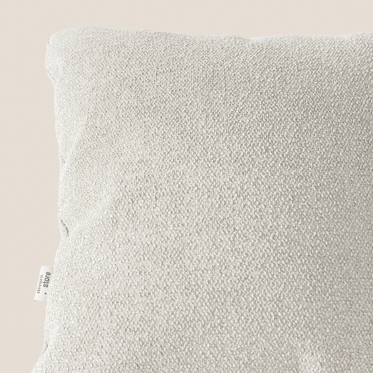 Primo piano del cuscino Lambada di divitt.sbs, in tessuto smacchiabile tipo cotone T33 grigio chiaro, su sfondo beige. La cucitura del cuscino presenta una piccola etichetta bianca con testo nero; è mostrata solo una parte del cuscino.