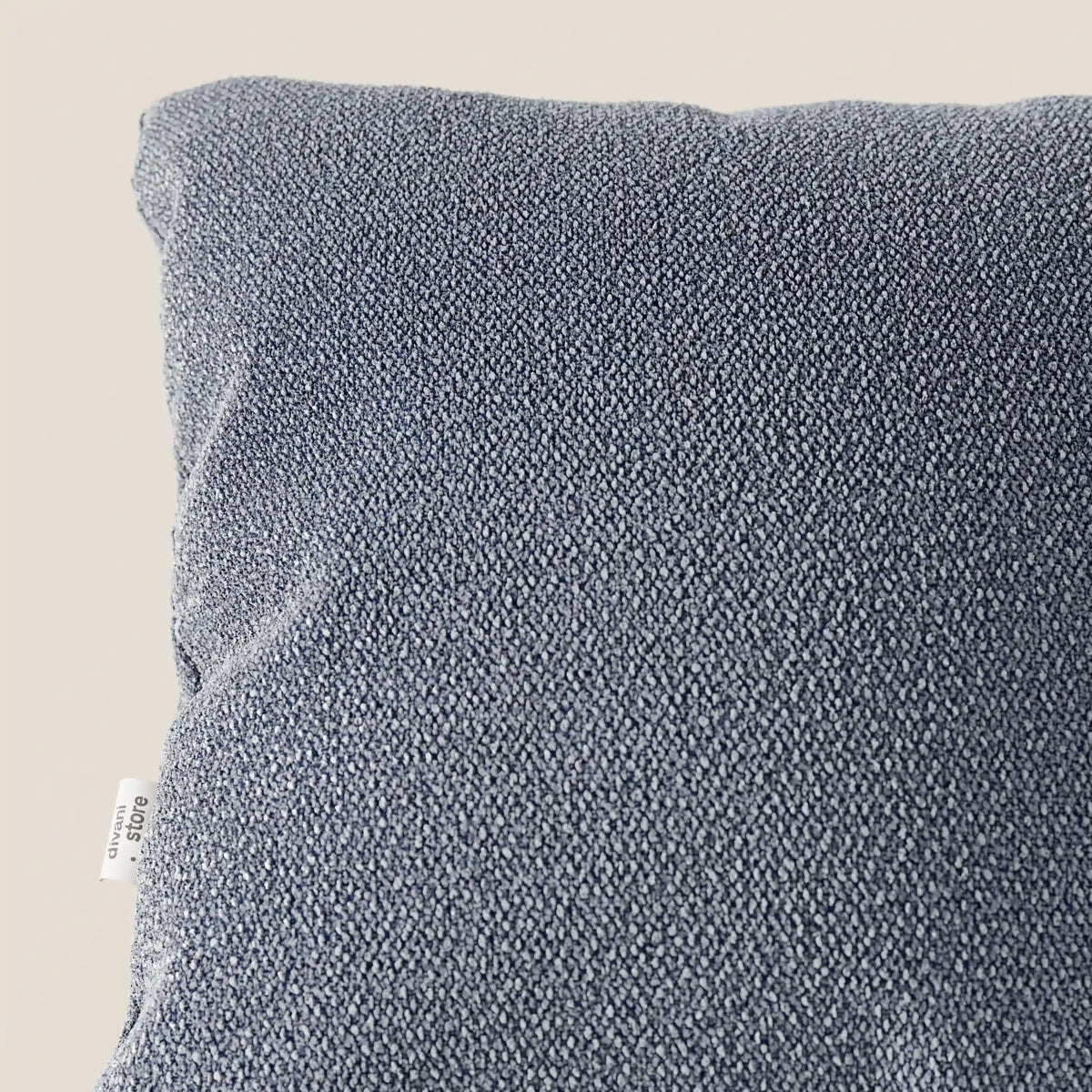 Primo piano del cuscino Lambada di divitt.sbs, in tessuto grigio-azzurro che sembra cotone naturale. Resistente alle macchie e all'acqua (T33), presenta una finitura intrecciata con una piccola etichetta bianca sul bordo sinistro.