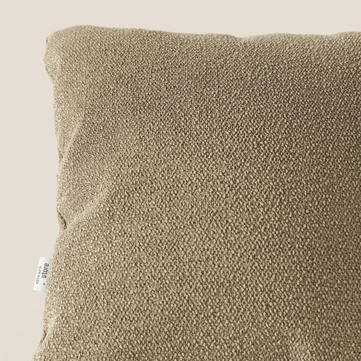 Primo piano dell'angolo superiore sinistro di un cuscino Lambada di divitt.sbs in color tortora, realizzato in tessuto naturale tipo cotone T33, fresco, antimacchia e impermeabile. Una piccola etichetta bianca con testo è visibile sulla cucitura sinistra; la superficie appare morbida e leggermente irregolare.