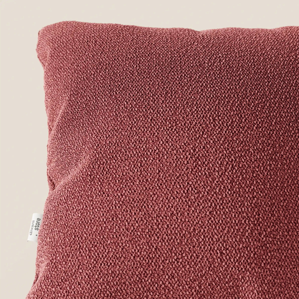 Primo piano del cuscino Lambada di divitt.sbs, in tessuto T33 rosso scuro, testurizzato, simile al cotone, smacchiabile e impermeabile, su sfondo beige. Una targhetta bianca con testo appare sul bordo sinistro del cuscino.