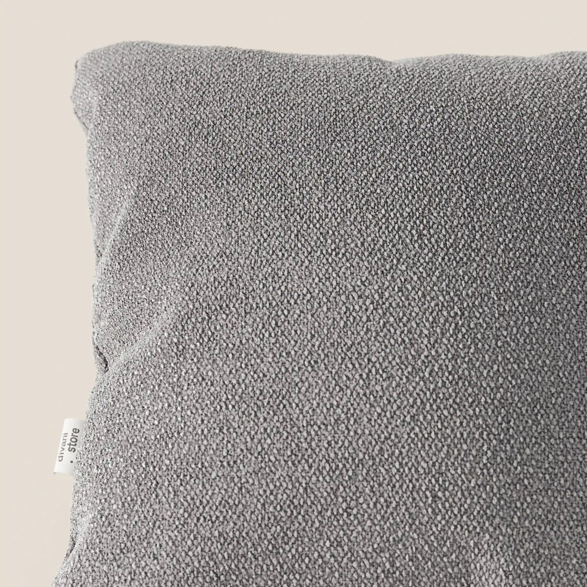 Primo piano dell'angolo superiore di un cuscino Lambada grigio di divitt.sbs, realizzato con un tessuto T33 fresco e naturale simile al cotone, resistente alle macchie e all'acqua. Una piccola etichetta bianca con testo appare sulla cucitura laterale vicino all'angolo.