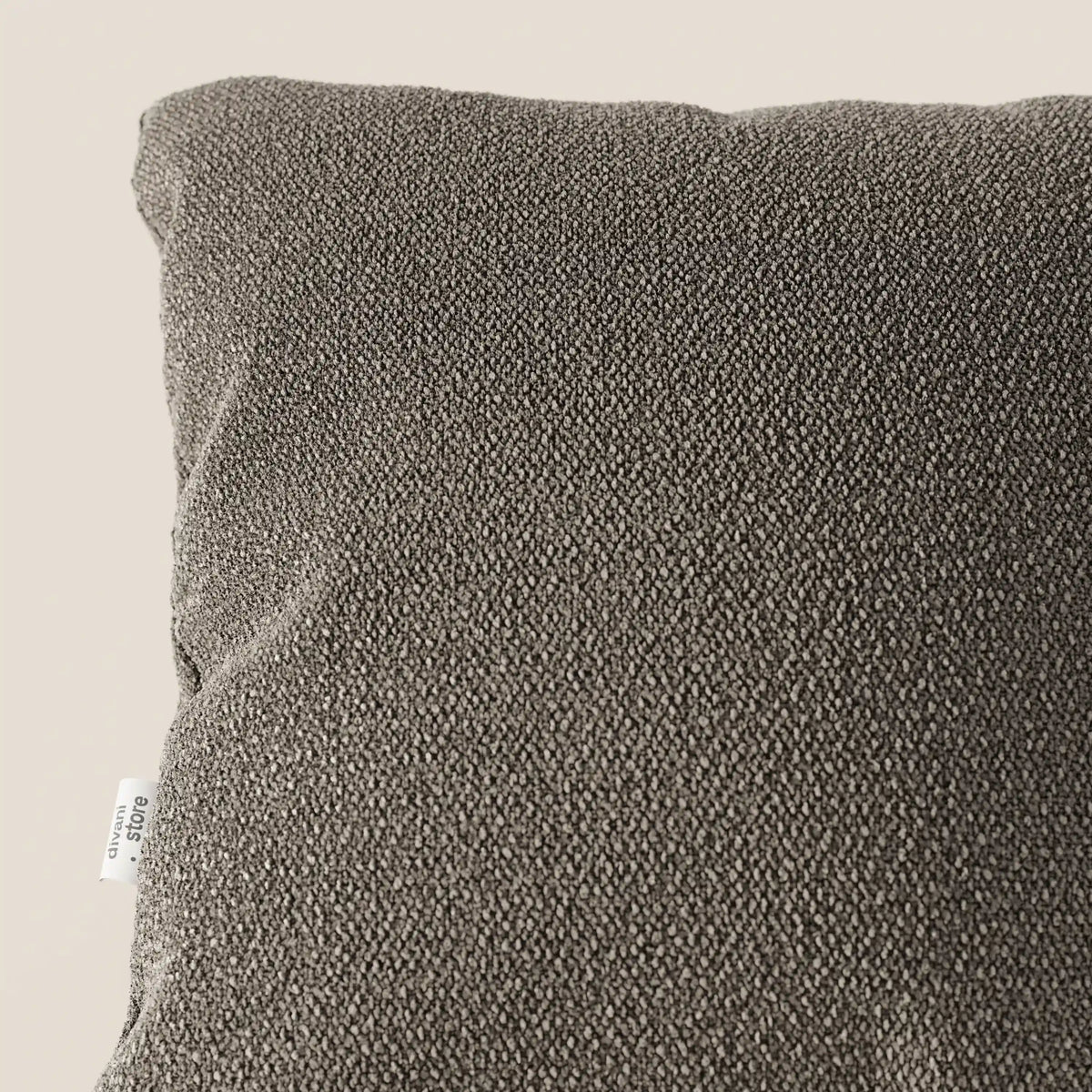 Primo piano dell'angolo superiore sinistro di un cuscino Lambada grigio scuro di divitt.sbs, realizzato in tessuto simil-cotone fresco, antimacchia e impermeabile (T33), con una piccola etichetta bianca sul bordo, su uno sfondo beige chiaro.