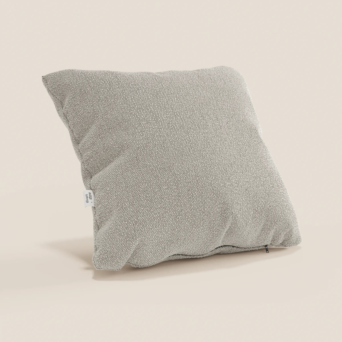 Un cuscino Lambada grigio chiaro di divitt.sbs in tessuto smacchiabile e impermeabile tipo cotone naturale si erge su uno sfondo beige. Su un lato è presente una piccola etichetta bianca e sul bordo inferiore è visibile una cerniera nera.