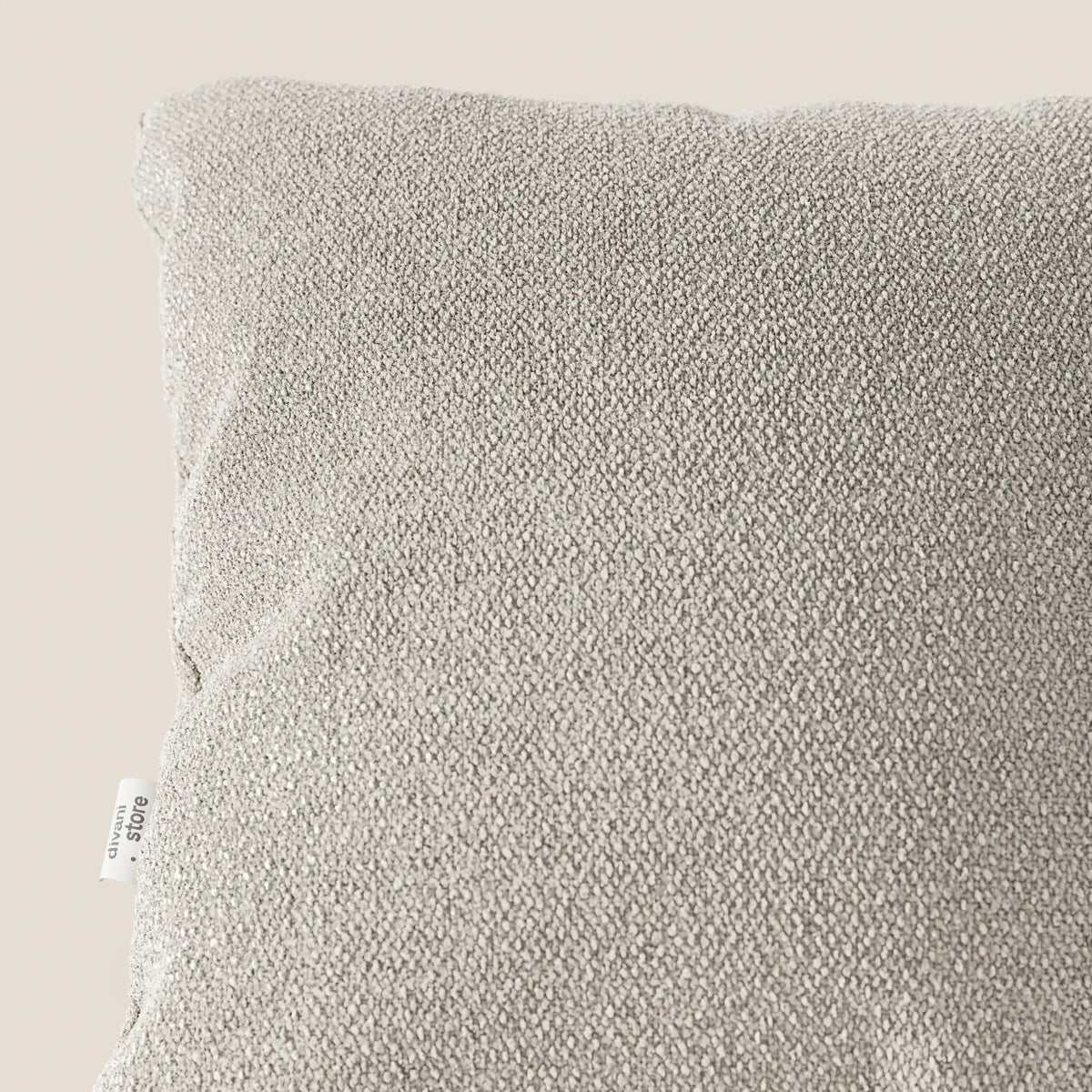 Primo piano di un cuscino Lambada grigio chiaro di divitt.sbs, realizzato in tessuto naturale simil-cotone fresco, antimacchia e impermeabile. Nell'angolo in alto a sinistra si nota un'etichetta bianca con testo nero sulla cucitura laterale su uno sfondo a tinta unita.