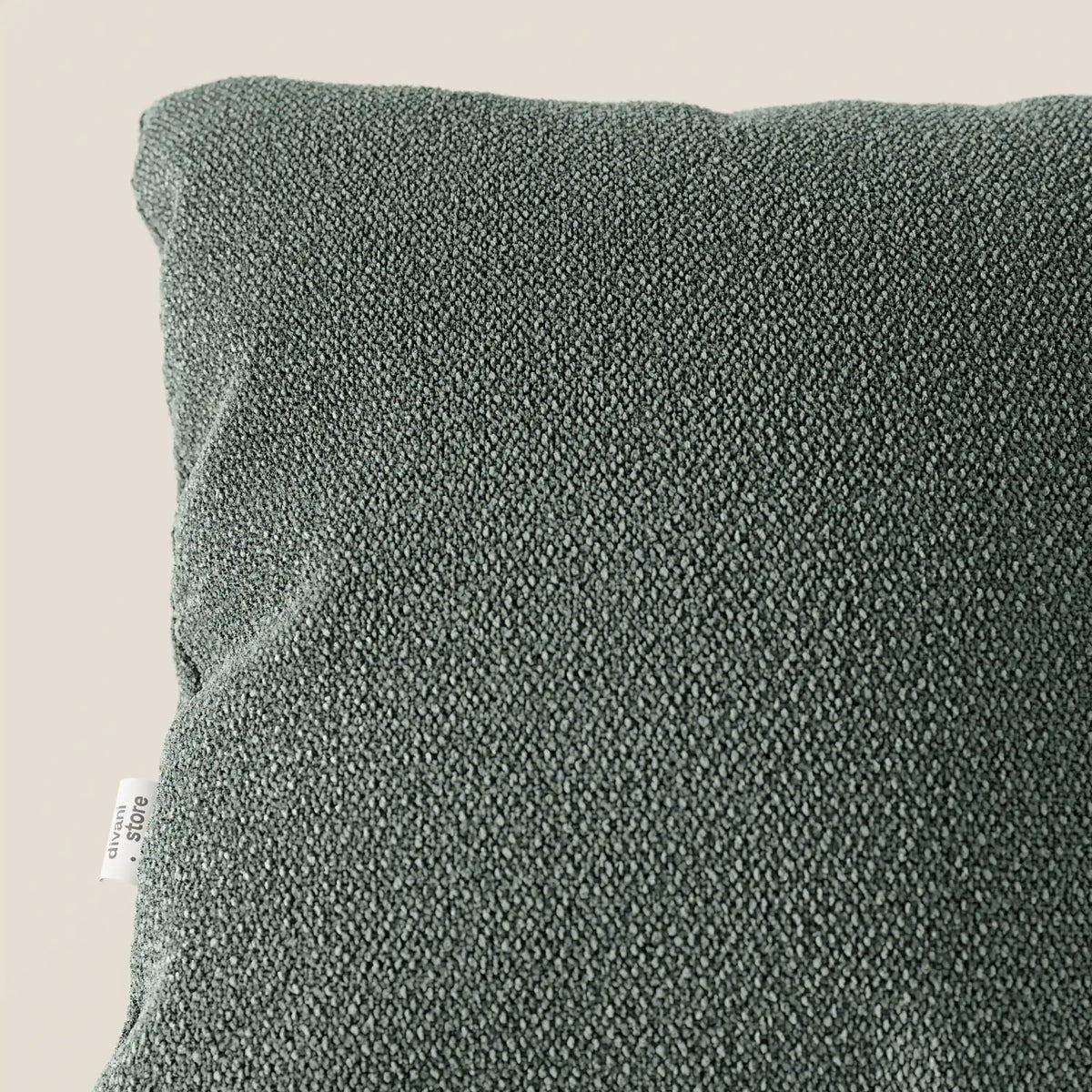 Primo piano del cuscino Lambada di divitt.sbs, di colore verde scuro, realizzato in tessuto fresco, antimacchia e impermeabile tipo cotone (T33), appoggiato su una superficie beige chiaro. Sul bordo sinistro compare una piccola etichetta bianca, visibile solo in parte.