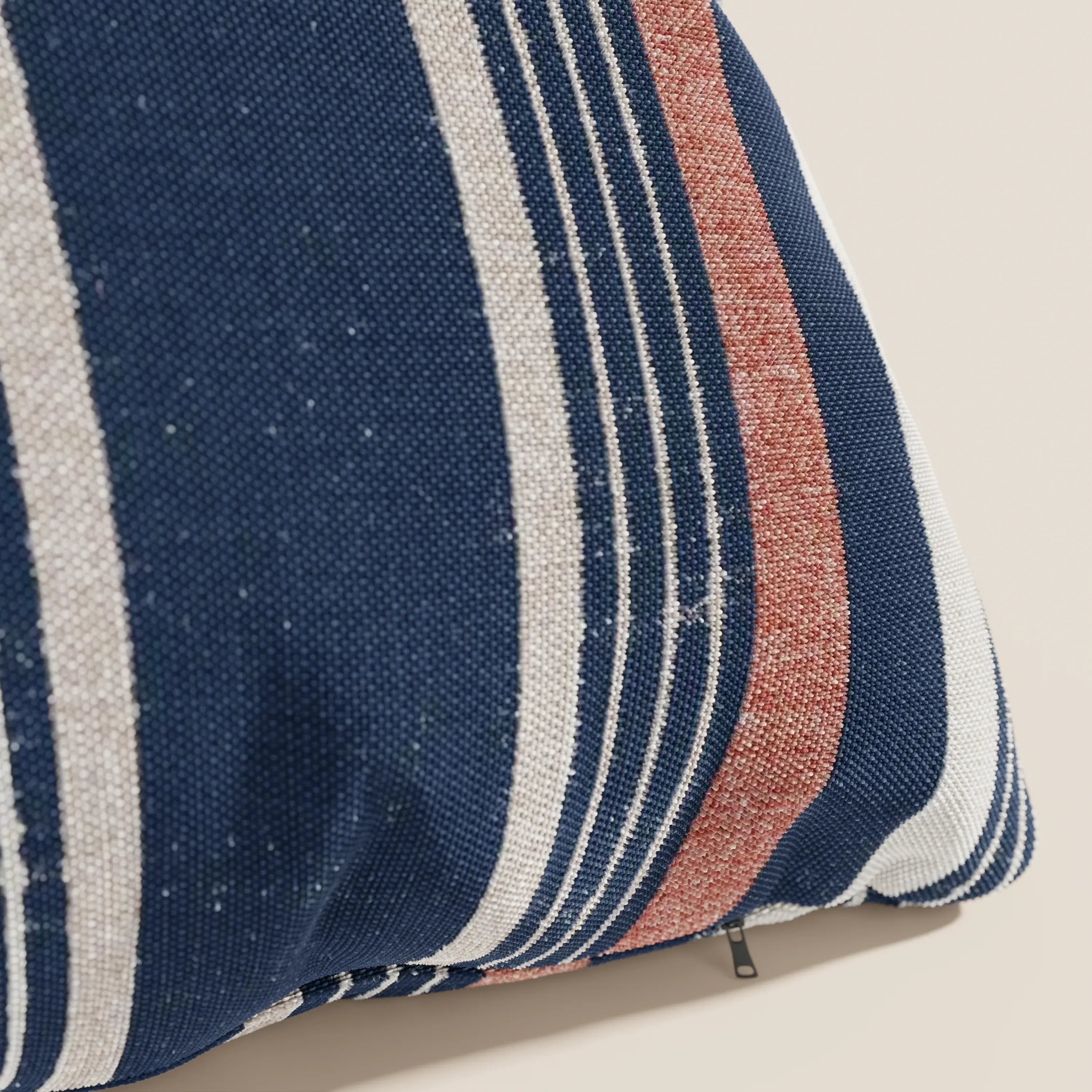 Primo piano di un cuscino a fantasia rigata 45x45 di divitt.sbs, in tessuto impermeabile arancio, caratterizzato da ampie strisce blu, bianche e rosso ruggine di spessore variabile. Una piccola cerniera è visibile su uno sfondo beige chiaro.