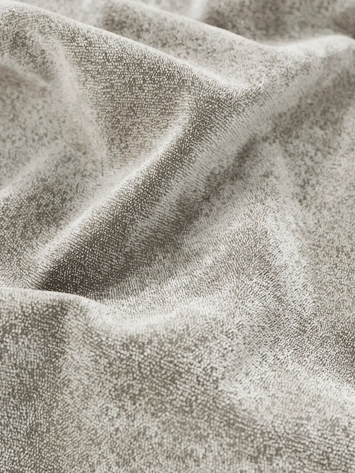 Un primo piano del tessuto in ciniglia grigio chiaro di un letto imbottito AURO con vano contenitore e testiera capitonné alta in ciniglia Oeko-Tex®, che mette in risalto la morbida trama drappeggiata in delicate pieghe che creano sottili giochi di luci e ombre.
