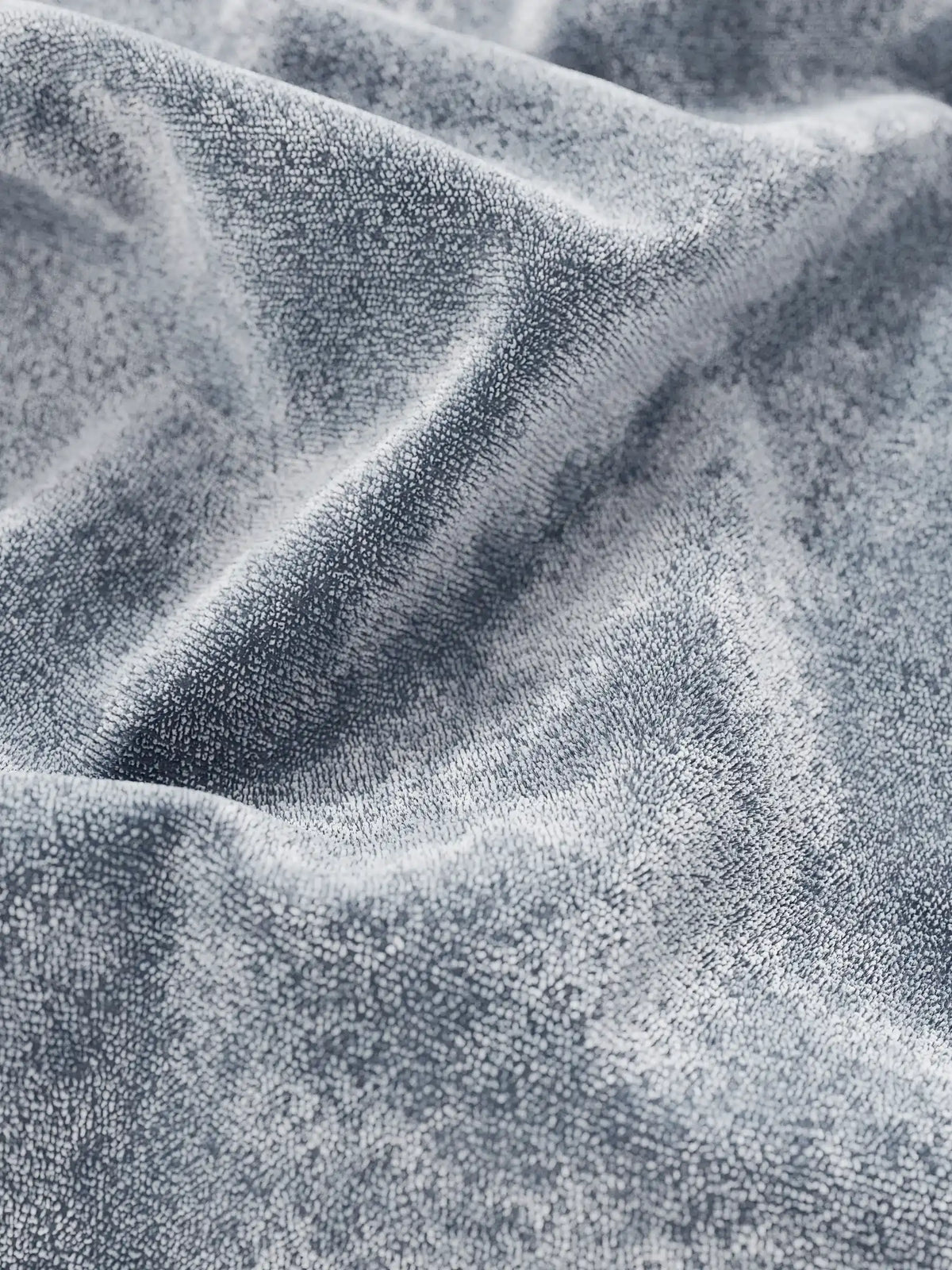 Primo piano su un morbido tessuto in ciniglia grigia dal pelo fitto e strutturato, drappeggiato in pieghe profonde sul letto imbottito AURO con vano contenitore e testiera capitonné alta in ciniglia Oeko-Tex®, creando un gioco di luci e ombre.