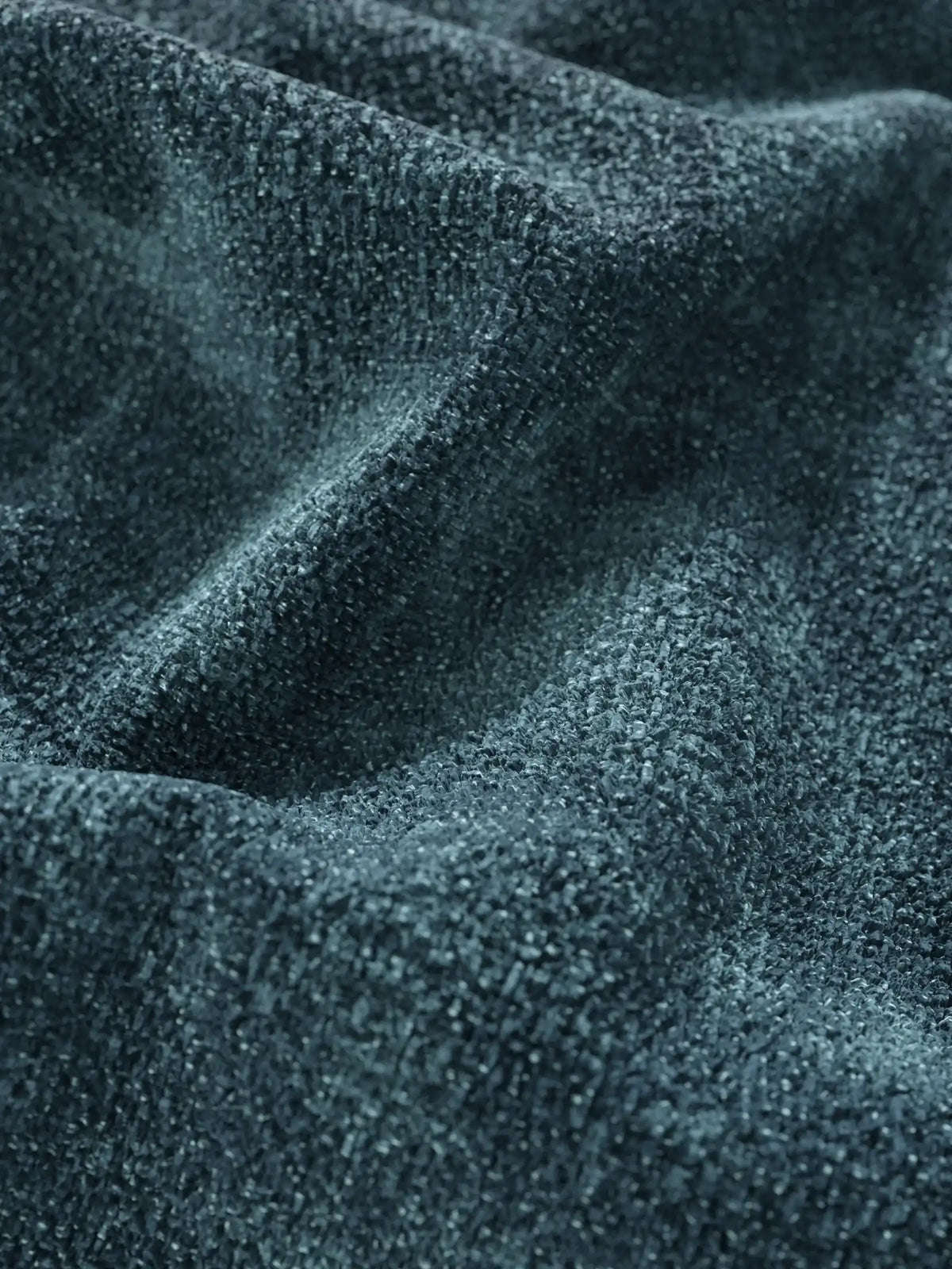 Primo piano su un tessuto color verde acqua scuro dalla trama ruvida, drappeggiato in morbide pieghe su un divano letto Ottaviano con letto estraibile – penisola reversibile e contenitore in tessuto materico lavabile, caratterizzato da una trama a grana grossolana con sottili variazioni di luce e ombra.