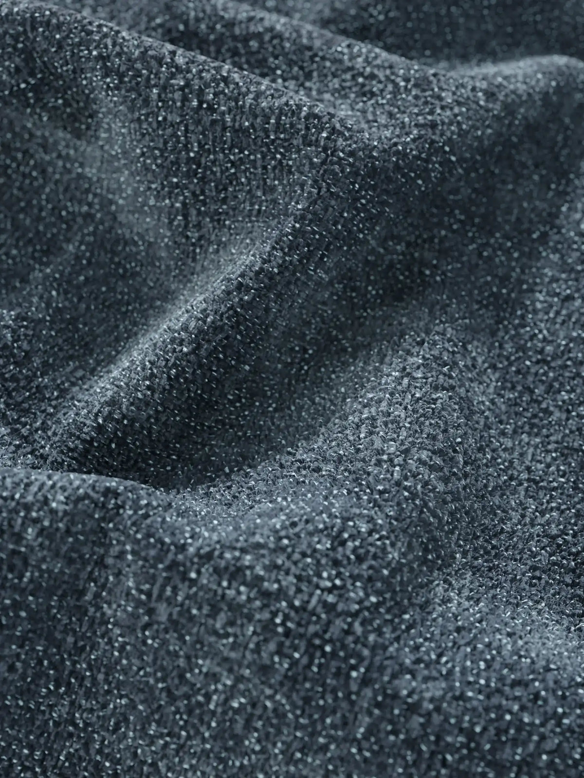 Un primo piano del tessuto grigio antracite su un divano letto con letto estraibile Ottaviano – penisola reversibile e contenitore in tessuto materico lavabile, con una trama luccicante che si raccoglie in morbide pieghe per mettere in risalto la trama intricata e granulosa.