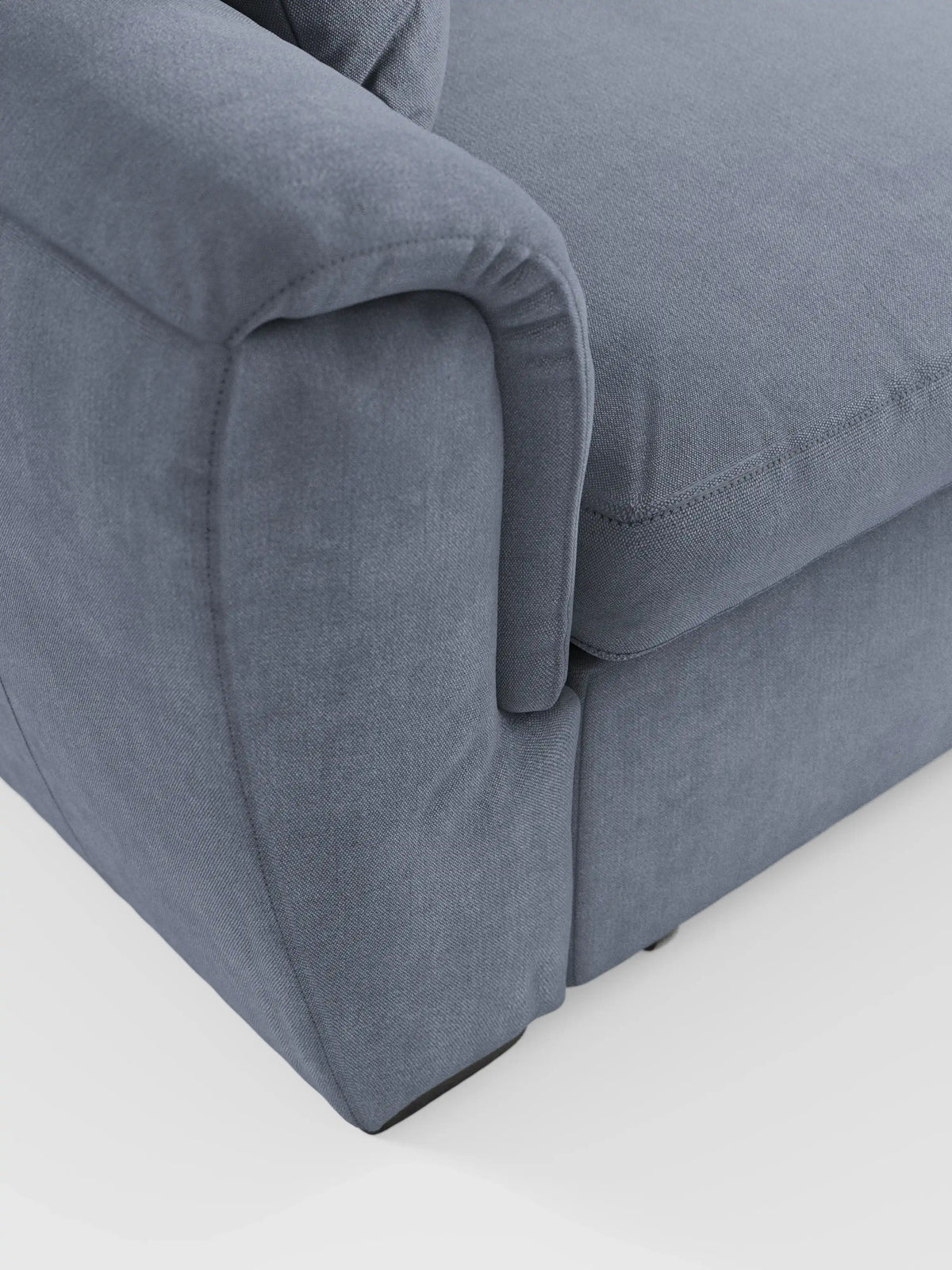 Primo piano del bracciolo e del cuscino di seduta del divano angolare reversibile con contenitore estraibile e pouf di divitt.sbs. Rivestito in morbido tessuto Smacchiabile Oeko-Tex® blu-grigio, il divano presenta cuciture a vista lungo i bordi. Il suo design mette in risalto i braccioli arrotondati e le linee morbide e pulite con poggiatesta regolabili, il tutto su uno sfondo bianco a tinta unita.
