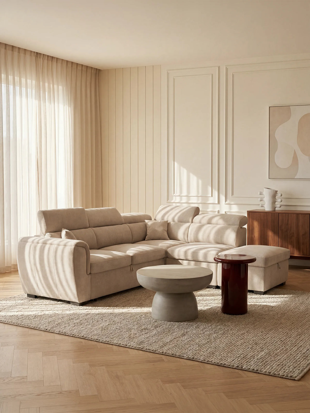 Un soggiorno moderno arredato con il divano angolare reversibile con letto estraibile e pouf contenitore Titano di divitt.sbs — un elegante divano componibile con poggiatesta reclinabili in tessuto antimacchia certificato Oeko-Tex® — è abbinato a un tappeto dalla trama particolare, un tavolino rotondo in cemento grigio, un piccolo tavolino rosso, una credenza in legno, opere d’arte astratte appese alle pareti rivestite di pannelli dai toni neutri e tende trasparenti che diffondono la luce del sole