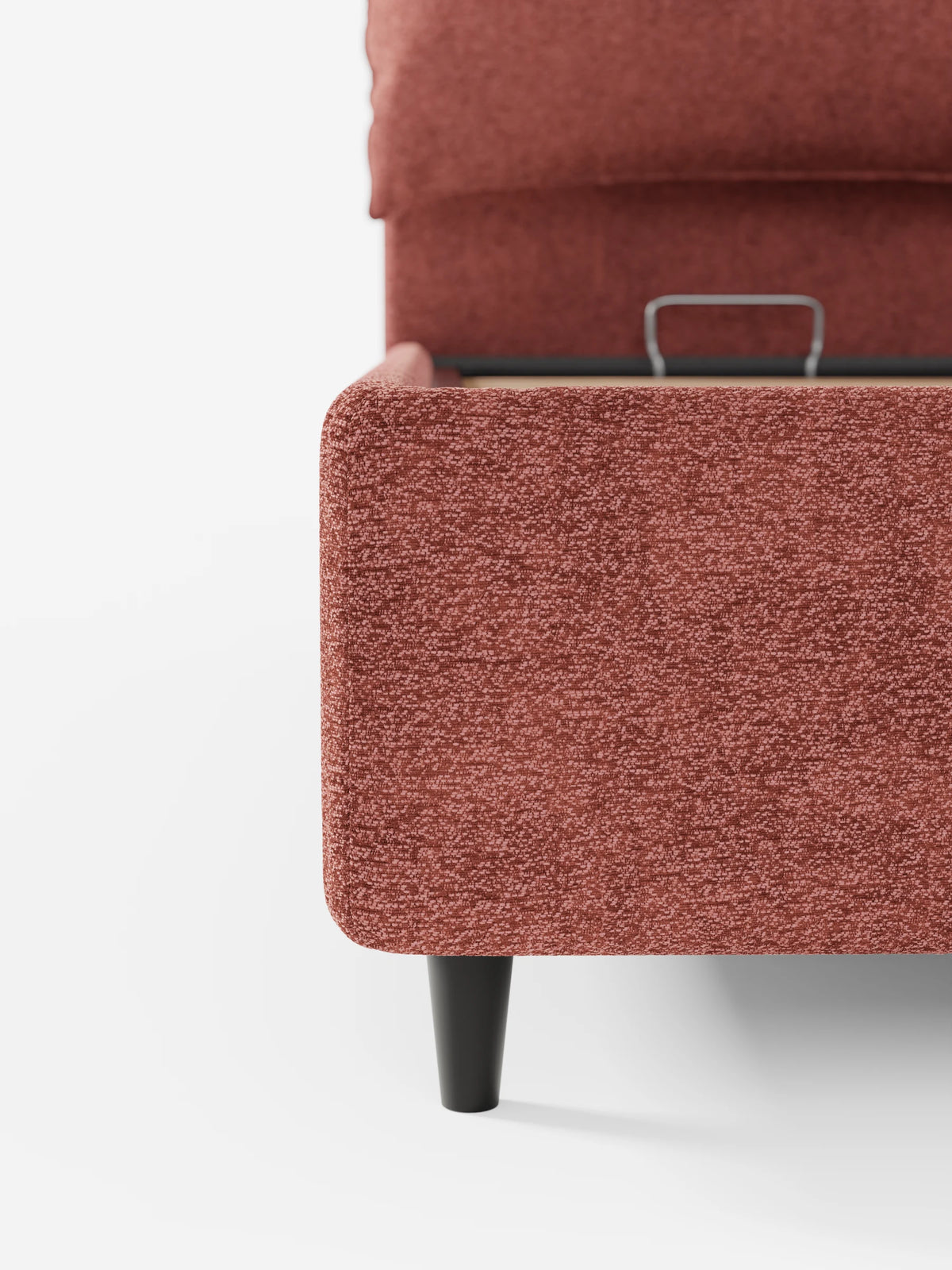 Primo piano di un letto imbottito ZAYRON color terracotta, dotato di vano contenitore e morbidi cuscini regolabili, poggiante su una gamba affusolata scura, con un vano contenitore parzialmente aperto visibile sotto la testiera regolabile.